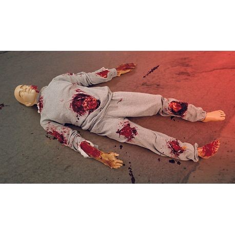 A realistic adult trauma and CPR manikin named Terry, featuring various simulated injuries like burns and lacerations, laid on a ground with emergency lights in the background.